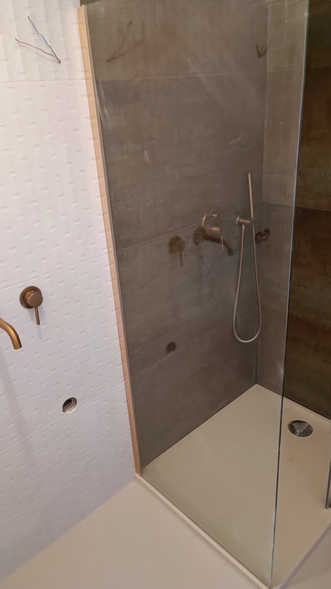 Niedokończona kabina prysznicowa z brązową armaturą, białe płytki z delikatnym wzorem na ścianie obok szarej ściany kabiny, widoczne przewody elektryczne wystające ze ściany.