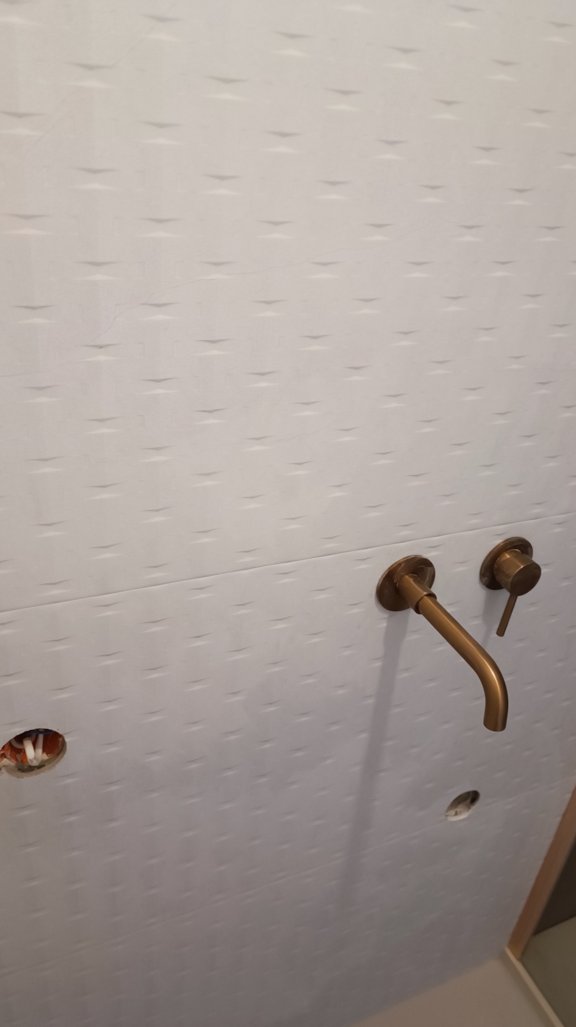 Ściana z białych, wzorzystych płytek ceramicznych, z zamontowaną baterią umywalkową w kolorze szczotkowanego złota. Widoczne otwory na rury i instalację elektryczną przed zakończeniem prac.