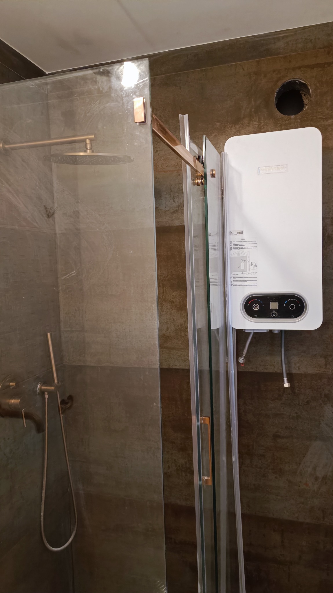 Nowoczesna kabina prysznicowa z elementami w kolorze miedzi, na ścianie zamontowany biały podgrzewacz wody, widoczne szare płytki imitujące beton.
