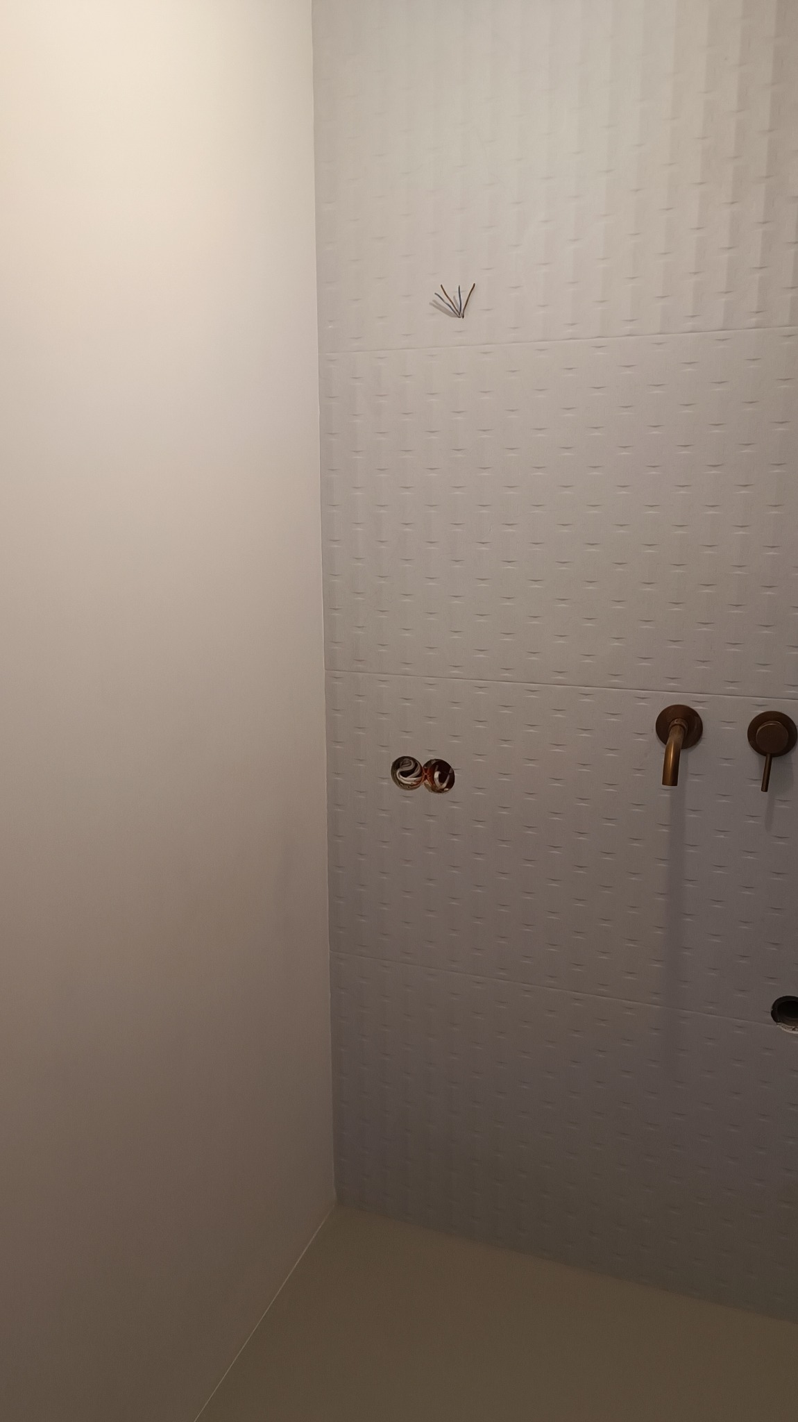 Ściana łazienki wyłożona białymi płytkami z delikatnym wzorem, widoczne wyprowadzenia rur i zamontowana bateria w kolorze mosiądzu, fragment ściany pomalowanej na biało oraz kilka zapałek...