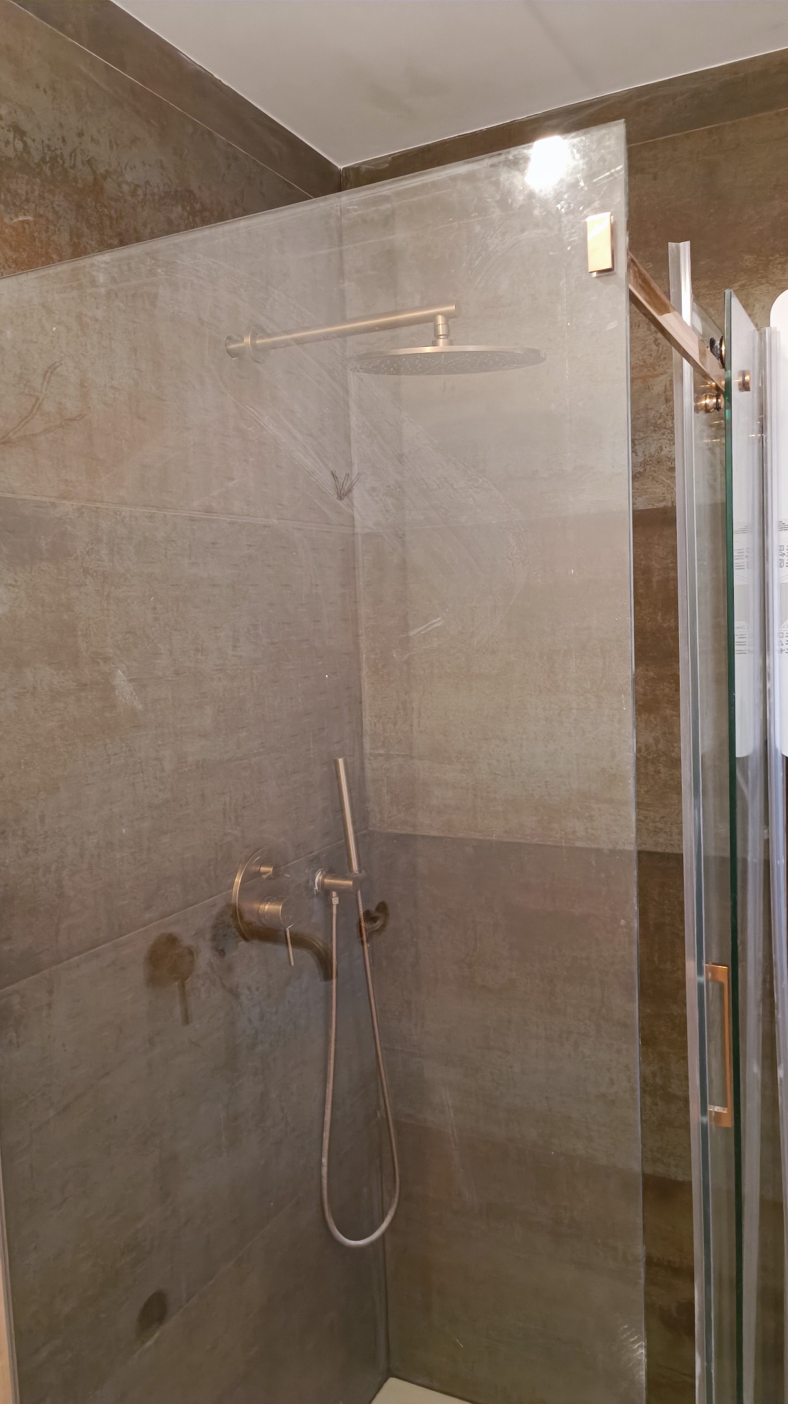 Nowoczesna kabina prysznicowa z deszczownicą i baterią w kolorze mosiądzu, wykończona płytkami imitującymi beton, widoczna przez lekko zabrudzoną szybę.