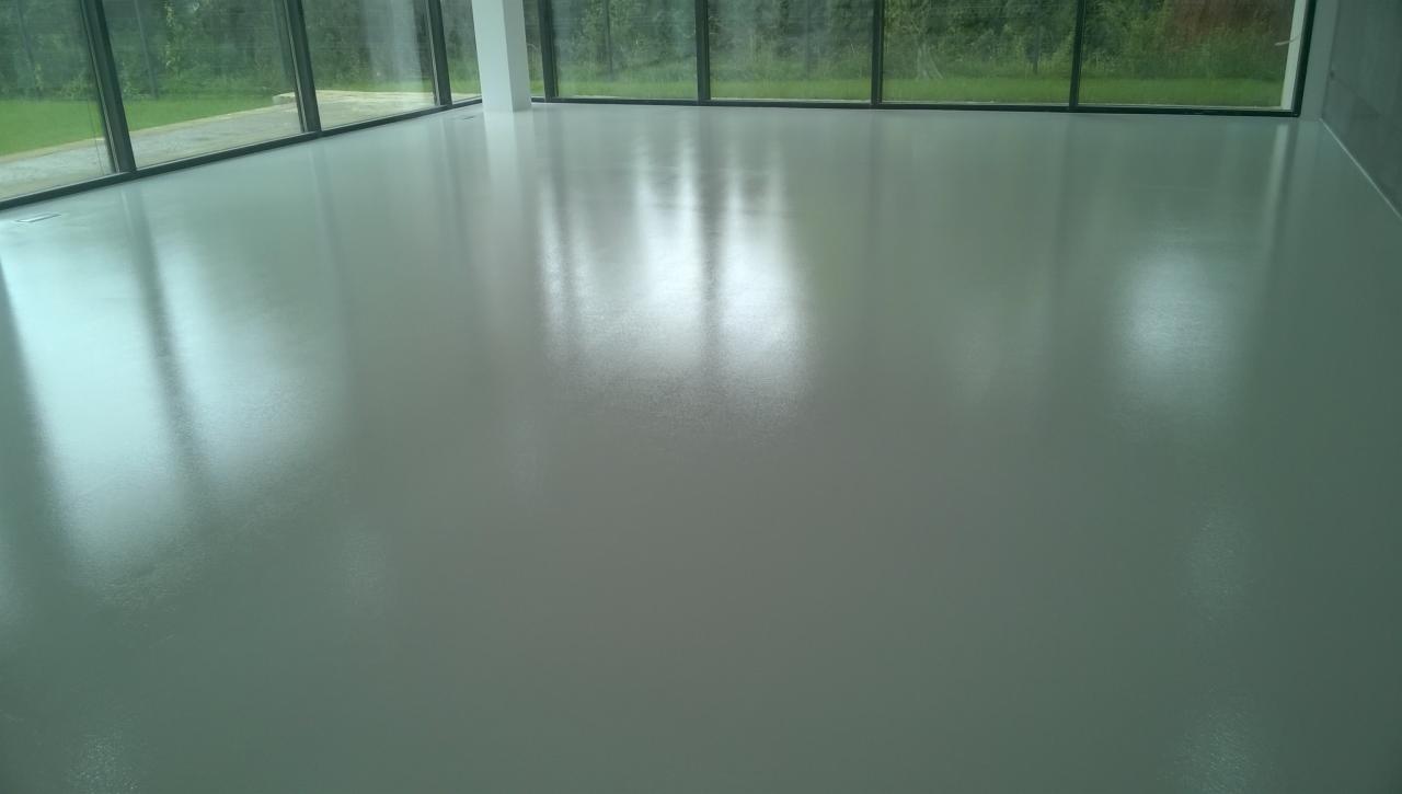 Jasnoszara, błyszcząca podłoga w przestronnym wnętrzu z dużymi oknami, odbijającymi zieleń ogrodu, minimalistyczny design.