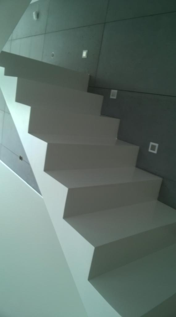 Nowoczesne, białe schody z minimalistyczną balustradą, przylegające do szarej ściany z kwadratowymi lampami.