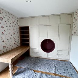 ModernCraft Norbert Ostrowski - Kremowa szafa z okrągłą wnęką i drewniane biurko z toczoną nogą, na tle kwiecistej tapety. Realizacja w Woli Dębińskiej. Nietypowe rozwiązanie do pokoju dziecięcego.