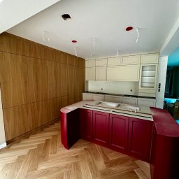 ModernCraft Norbert Ostrowski - Montaż kuchni: bordowa wyspa z blatem, beżowe szafki wiszące i drewniana zabudowa ściany. Parkiet w jodełkę, sufit z otworami na oświetlenie.