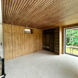 ModernCraft Norbert Ostrowski - Wnętrze z drewnianą zabudową: szafy wnękowe z jasnego drewna pod sufitem z drewnianych listew. Duże okno z widokiem na zieleń. Surowy beton na podłodze.
