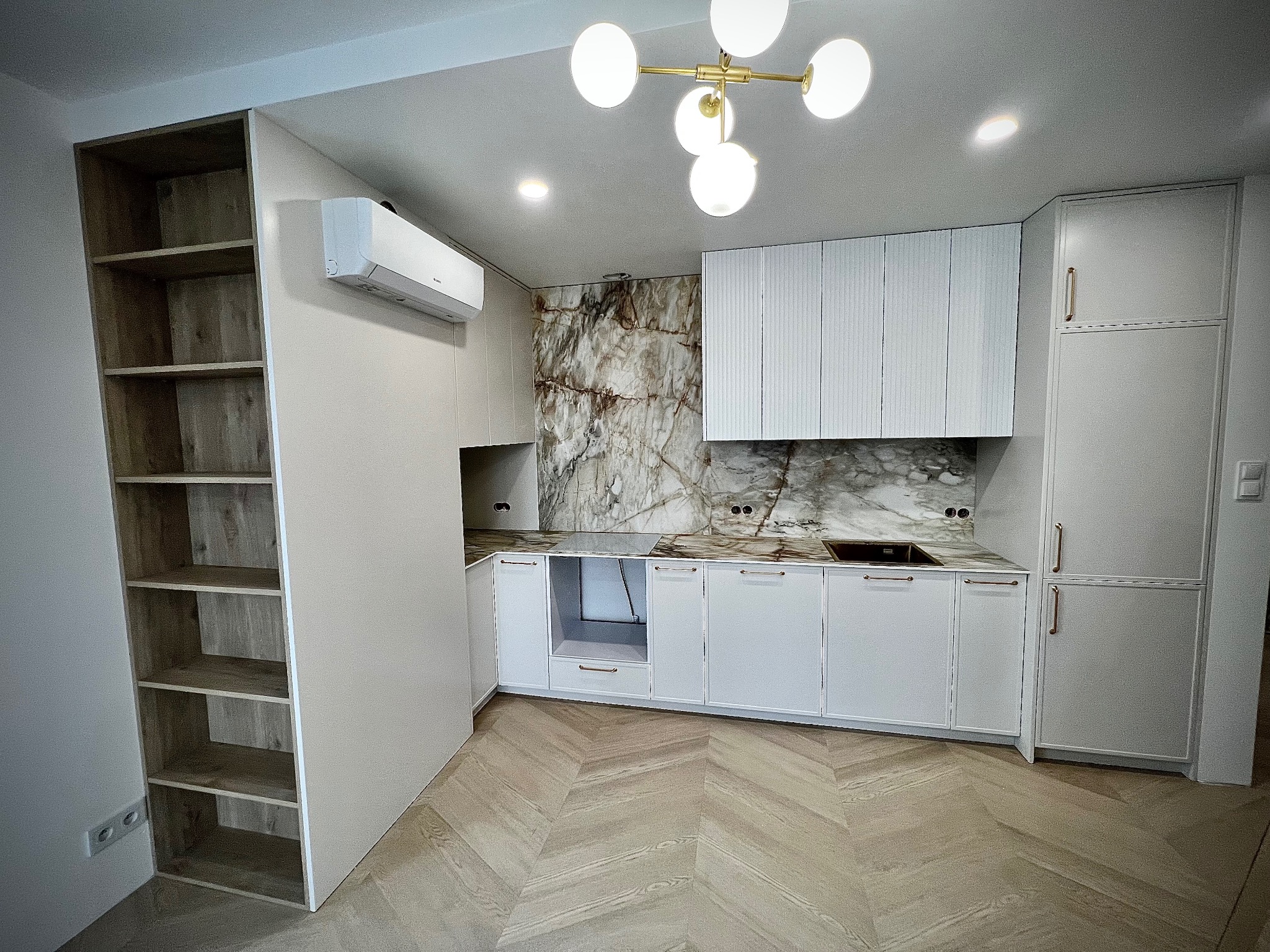 Nowoczesna kuchnia z białymi frontami, marmurowym blatem i ścianą. Oświetlenie punktowe i drewniana podłoga w jodełkę. Widoczny regał z półkami.