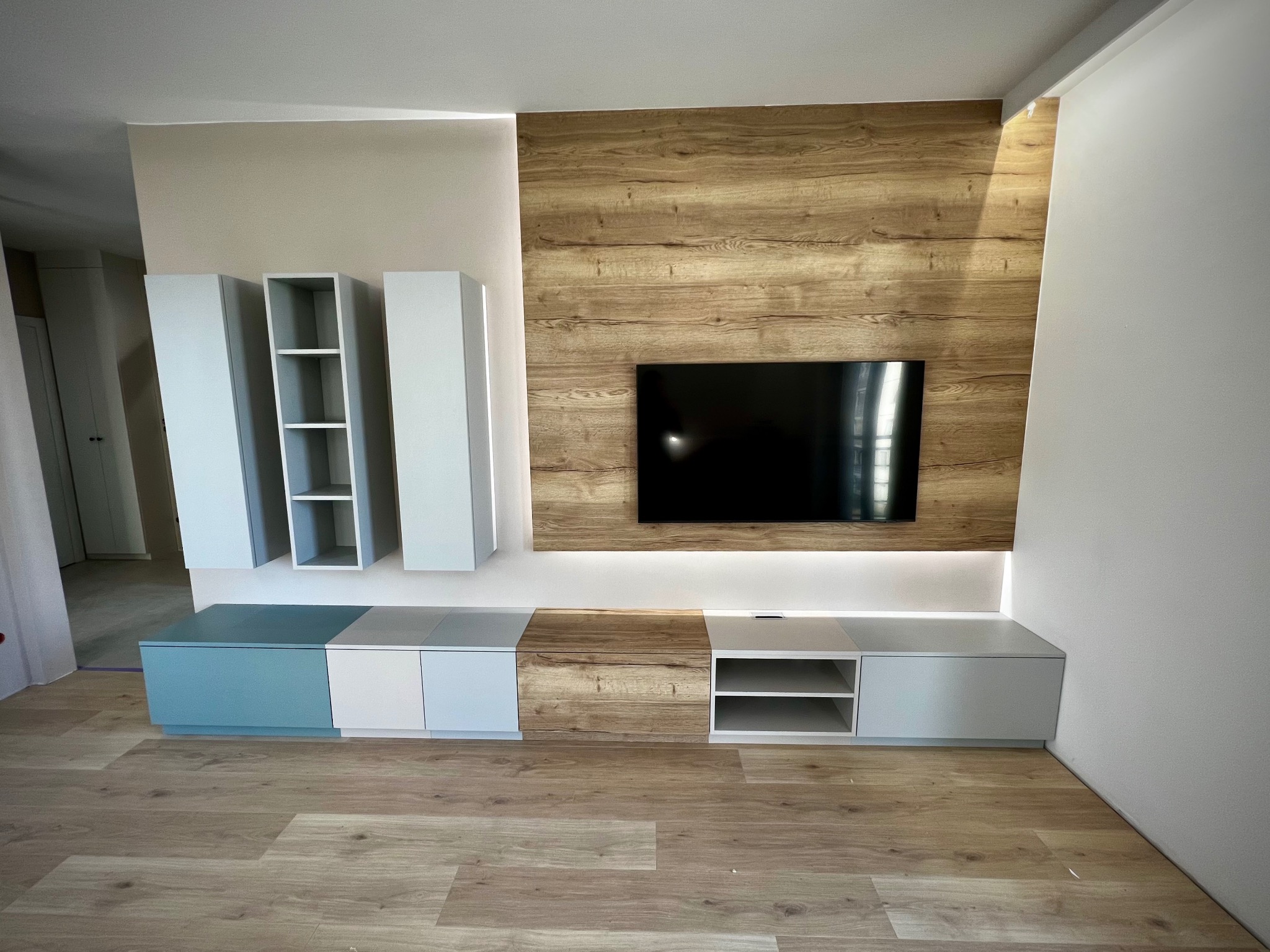 Nowoczesna zabudowa ściany TV z drewnianym panelem i podświetleniem LED. Szafki wiszące i stojące w odcieniach bieli, szarości i błękitu. Parkiet w kolorze jasnego drewna.