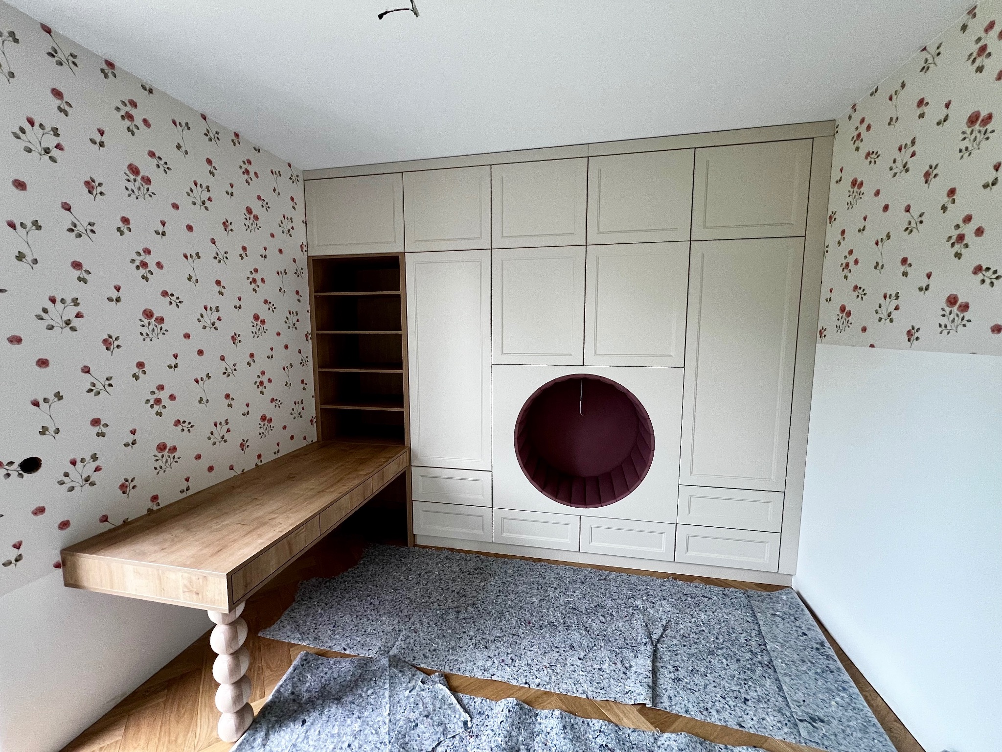 Kremowa szafa z okrągłą wnęką i drewniane biurko z toczoną nogą, na tle kwiecistej tapety. Realizacja w Woli Dębińskiej. Nietypowe rozwiązanie do pokoju dziecięcego.