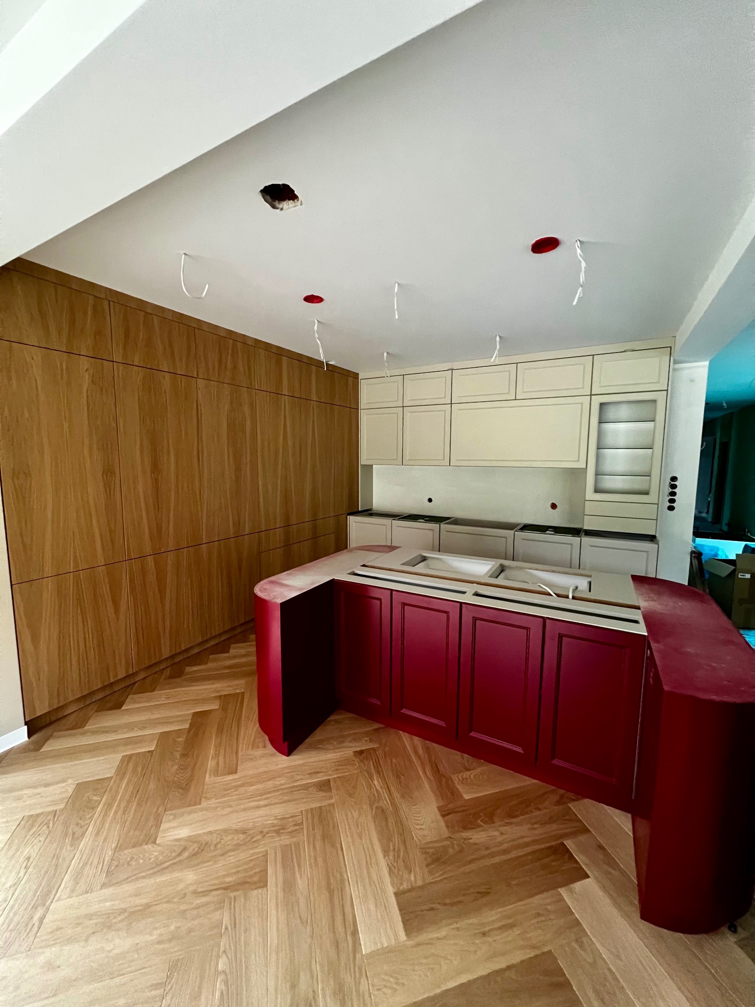 Montaż kuchni: bordowa wyspa z blatem, beżowe szafki wiszące i drewniana zabudowa ściany. Parkiet w jodełkę, sufit z otworami na oświetlenie.