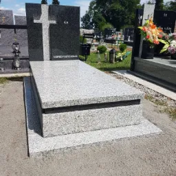 Granitowy grobowiec z krzyżem i napisem 'Na zawsze w naszych sercach', otoczony innymi nagrobkami na cmentarzu w słoneczny dzień.