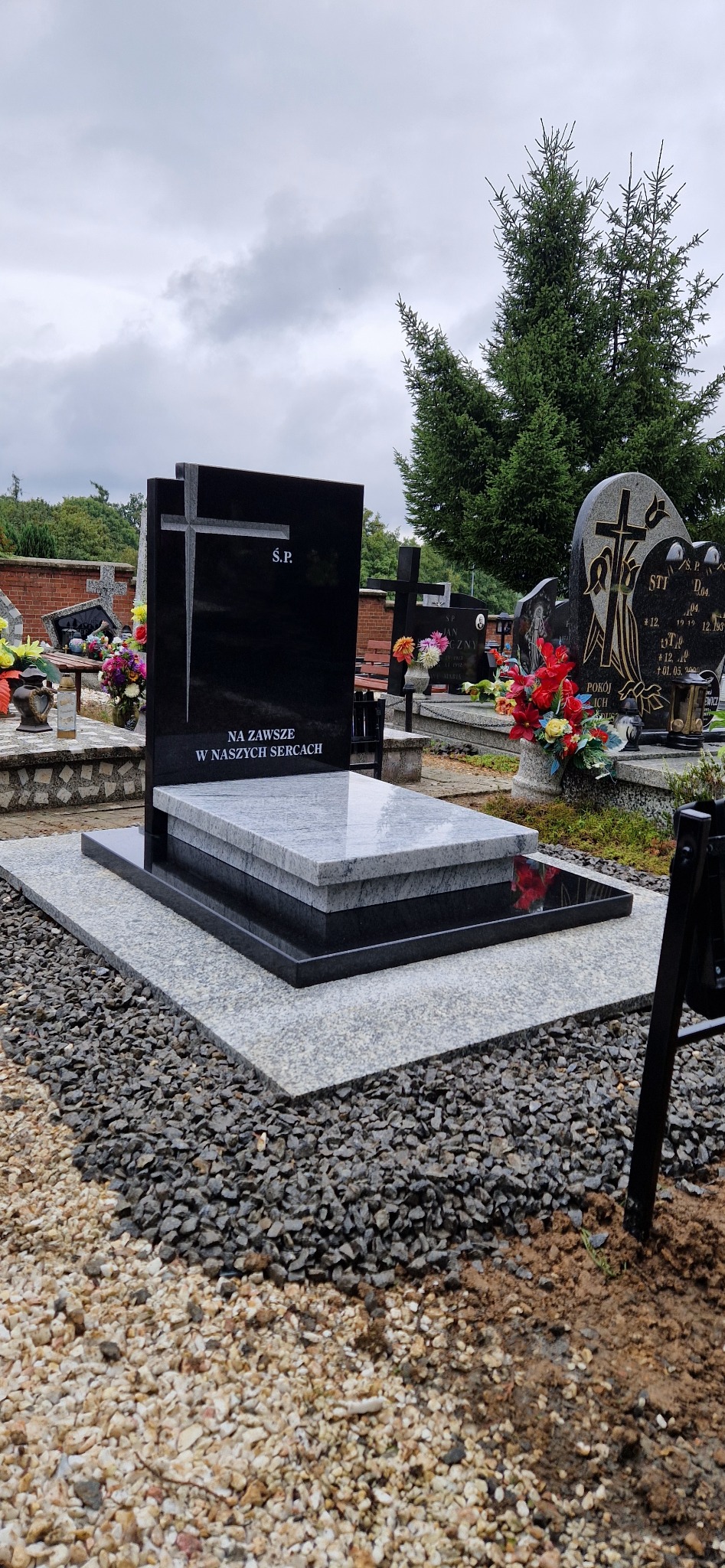 Czarny, nowoczesny pomnik z krzyżem i napisem 'Ś.P. Na zawsze w naszych sercach', otoczony szarym kamieniem na cmentarzu. Widok z bliska, pochmurne niebo.