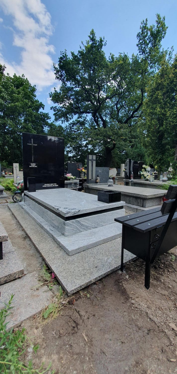 Czarny nagrobek z krzyżem na cmentarzu, otoczony zielenią drzew. Jasnoszary kamień podstawy i płyty nagrobnej. W tle inne groby i błękitne niebo.