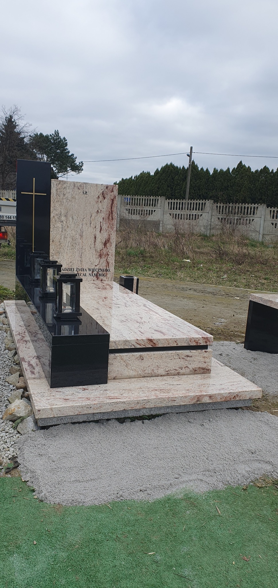 Granitowy pomnik z czarnym krzyżem i lampionami, jasny kamień z czerwonymi żyłkami, napis 'Nadziei życia wiecznego'. Tło: szare niebo, drzewa, płot.
