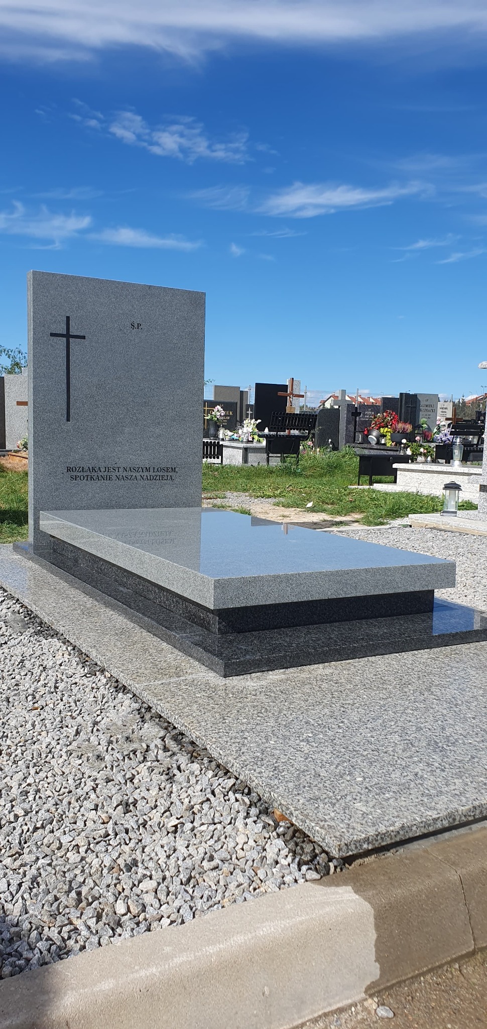 Szary, granitowy nagrobek z krzyżem i napisem na cmentarzu, otoczony żwirem. Błękitne niebo w tle. Polerowana powierzchnia płyty odbija światło.