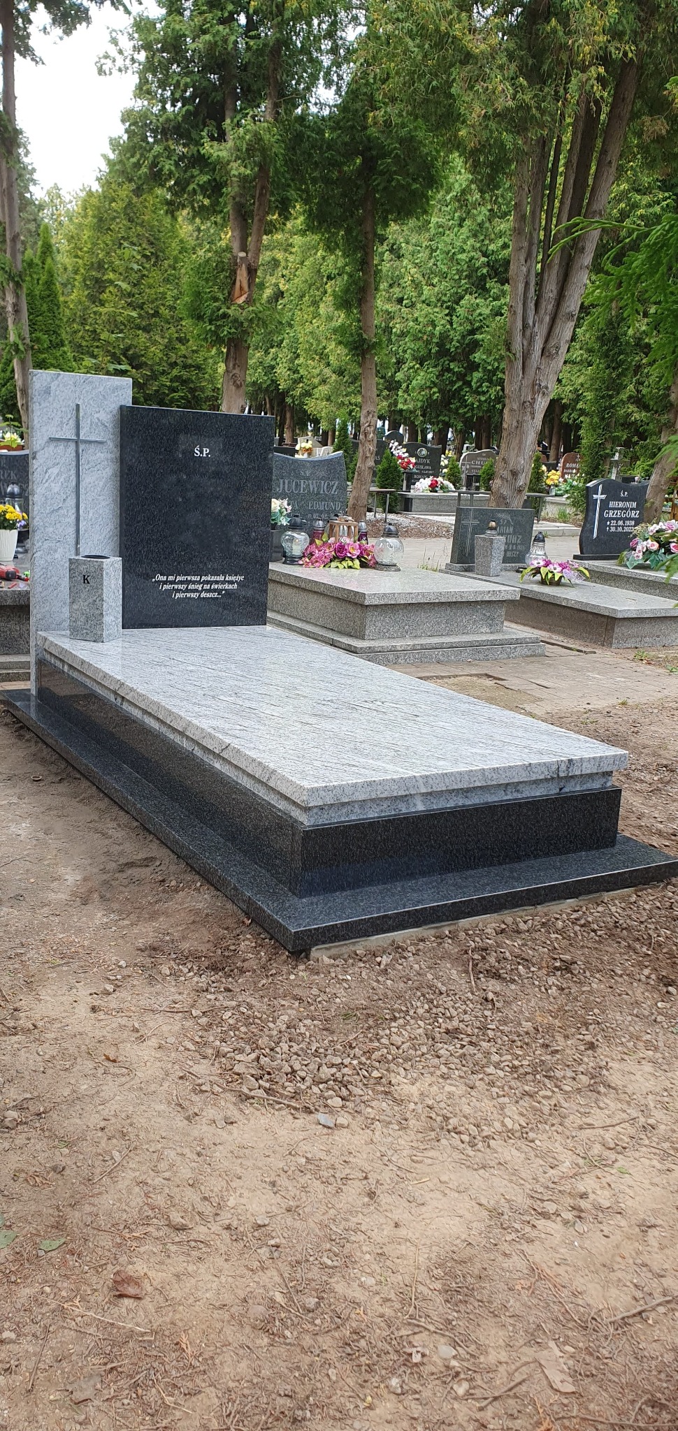 Dwukolorowy, granitowy pomnik z krzyżem i inskrypcją 'S.P.' na cmentarzu, otoczony zielenią drzew. Jasnoszary blat i ciemna podstawa. Widok z perspektywy ziemi.
