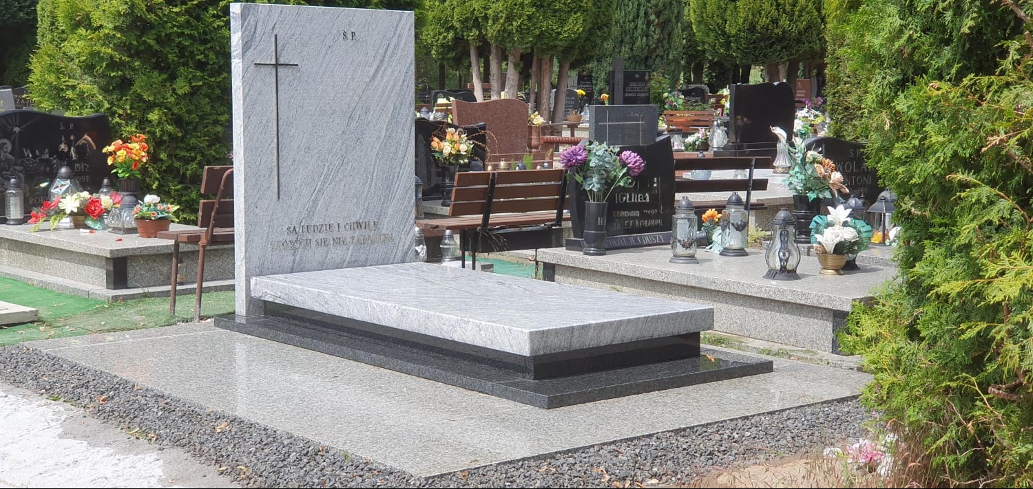Jasnoszary, granitowy pomnik z krzyżem na cmentarzu, otoczony zielenią drzew i krzewów. Widoczne inne groby w tle. Ujęcie w słoneczny dzień.