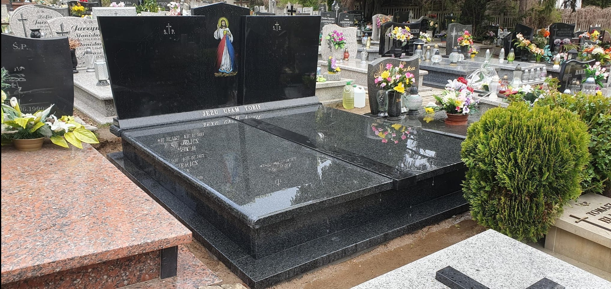 Czarny, podwójny pomnik z granitu z wizerunkiem Jezusa i napisem 'Jezu ufam Tobie' na cmentarzu, otoczony innymi nagrobkami i zielenią. Błyszcząca powierzchnia.