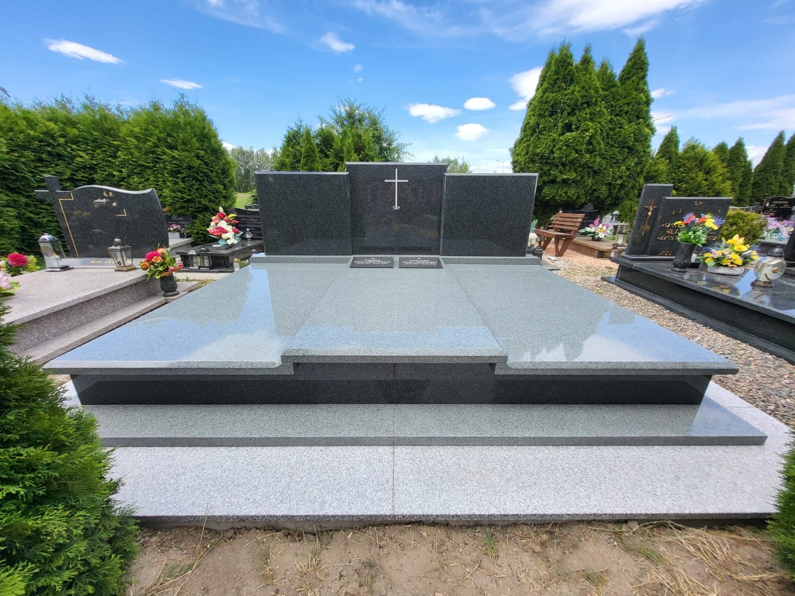 Nowoczesny, trzyczęściowy pomnik z ciemnego granitu z krzyżem, otoczony zielenią cmentarza i błękitnym niebem. Widoczne inne groby w tle.