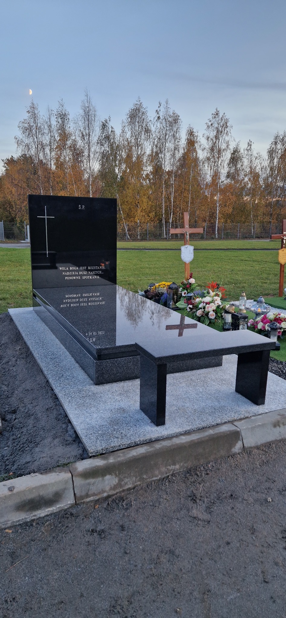 Czarny, nowoczesny pomnik z granitu z ławką na cmentarzu, w tle drzewa i niebo. Widoczny krzyż i inskrypcje. Minimalistyczny design.