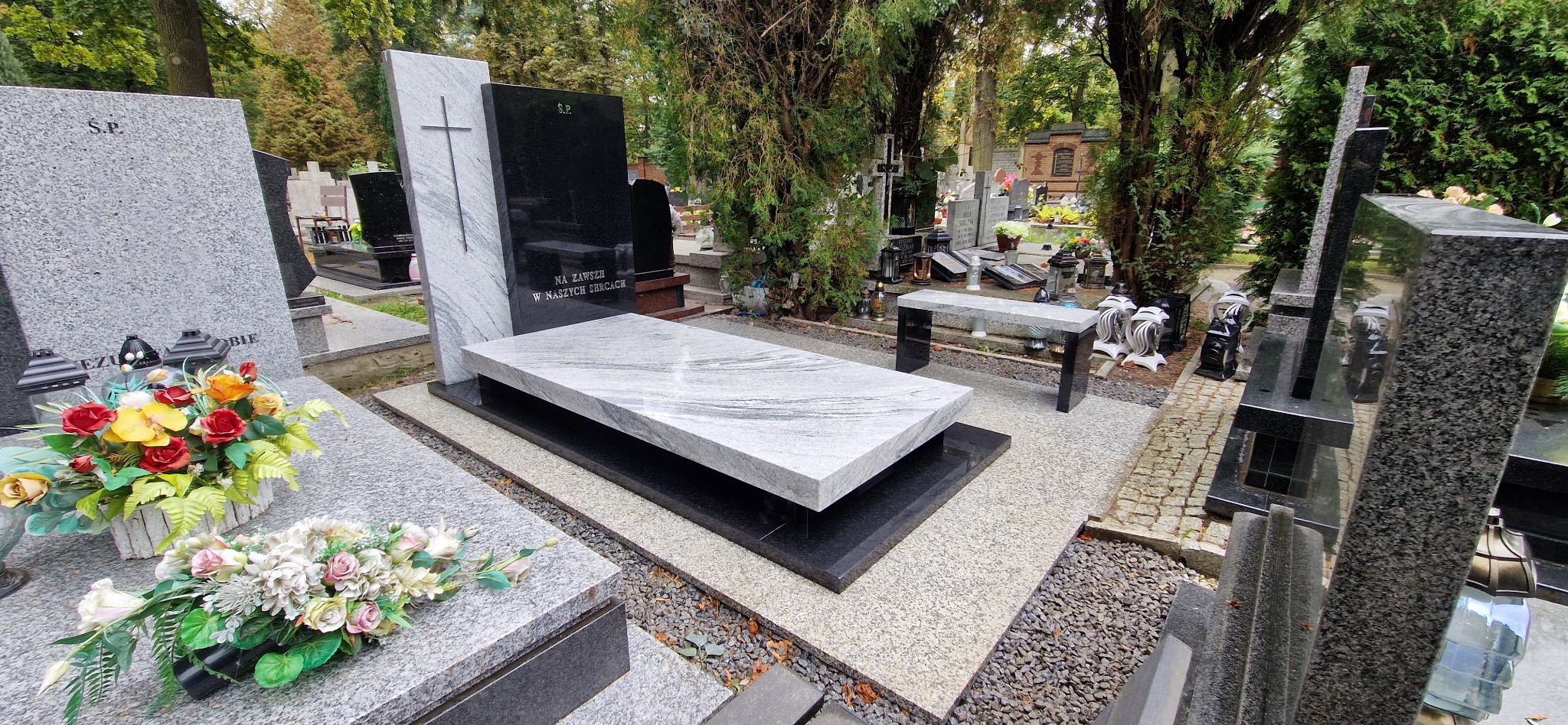 Nowoczesny nagrobek z jasnego i czarnego granitu na cmentarzu, z minimalistycznym krzyżem i ławką. Widok na inne groby i drzewa w tle.