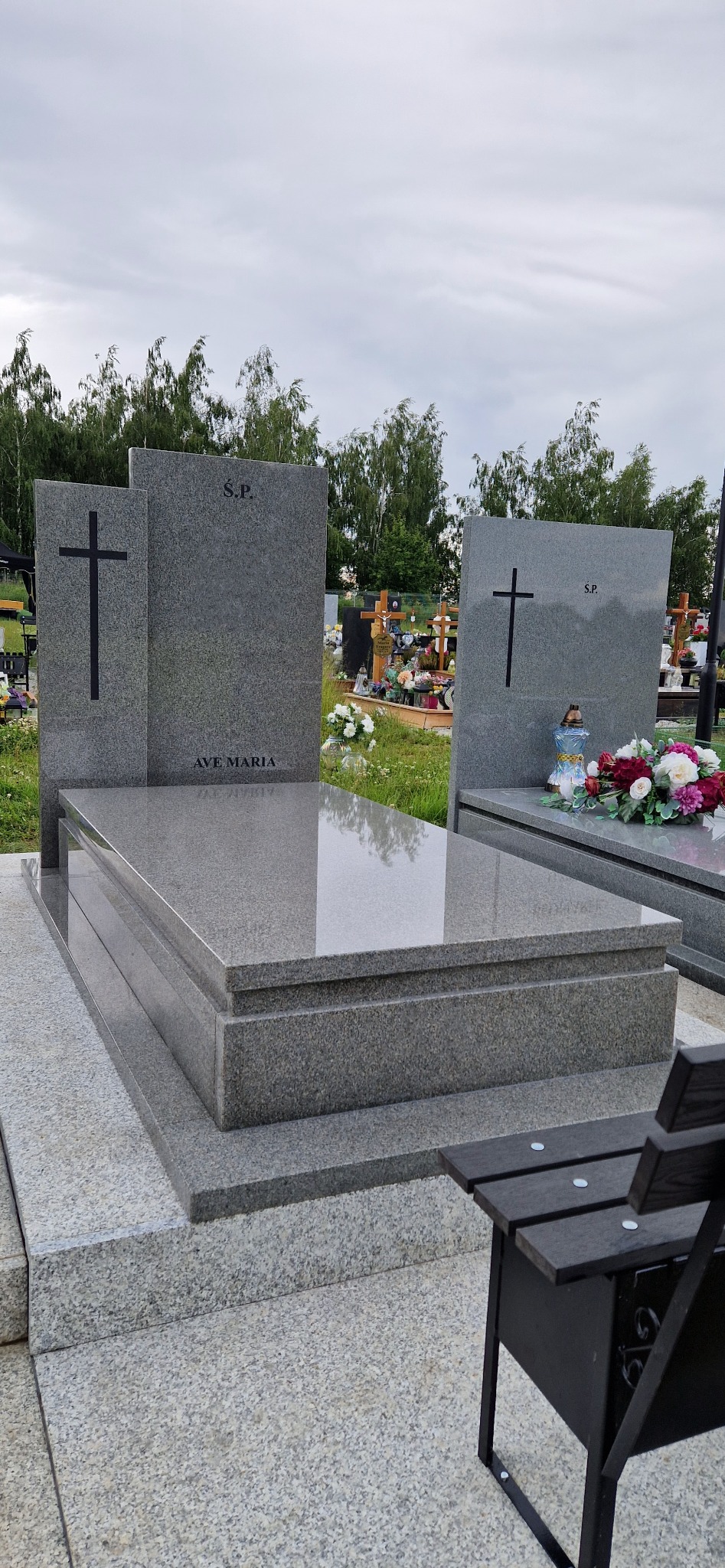 Szary, granitowy pomnik z napisem 'Ave Maria' i krzyżem, na cmentarzu, z ławką w prawym dolnym rogu. Widok na nagrobki i drzewa w tle, pochmurne niebo.