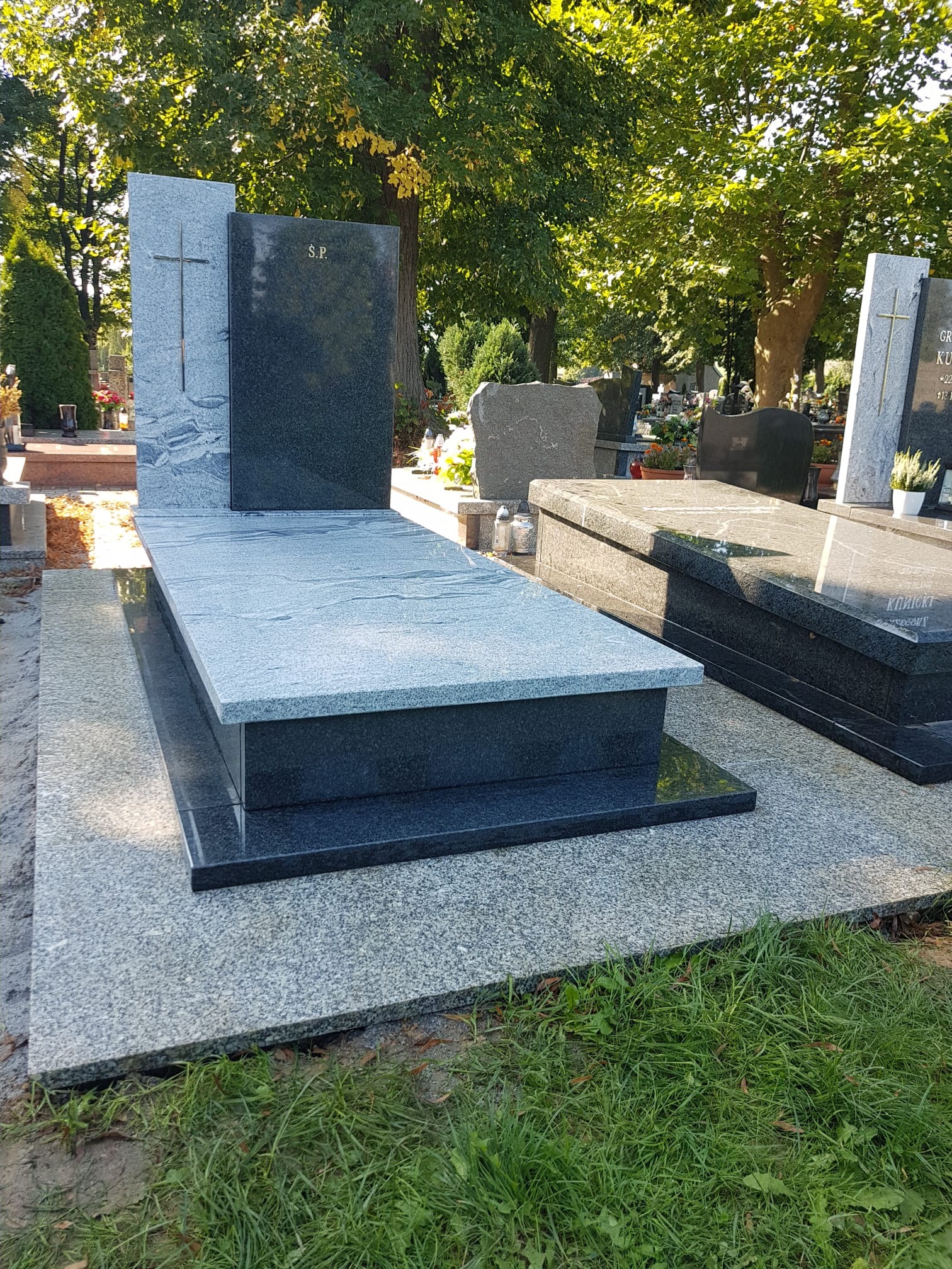 Nowoczesny, dwukolorowy pomnik z granitu, z wyrytym krzyżem i inicjałami 'S.P.', na cmentarzu, w otoczeniu zieleni drzew.
