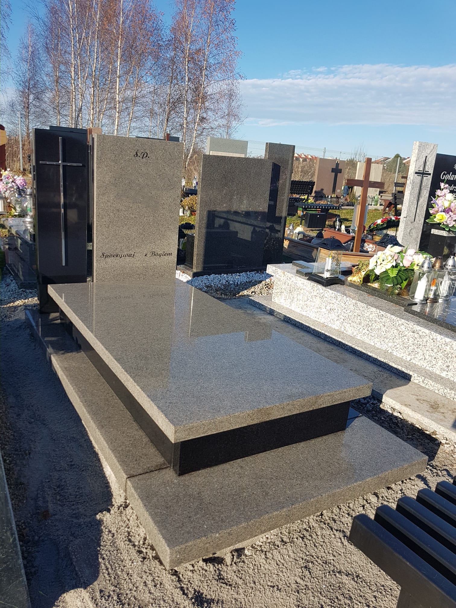 Nowoczesny, dwuelementowy pomnik z szarego granitu z wygrawerowanym napisem 'S.P. Spoczywaj z Bogiem', na tle cmentarza z innymi grobami i drzewami bez liści.