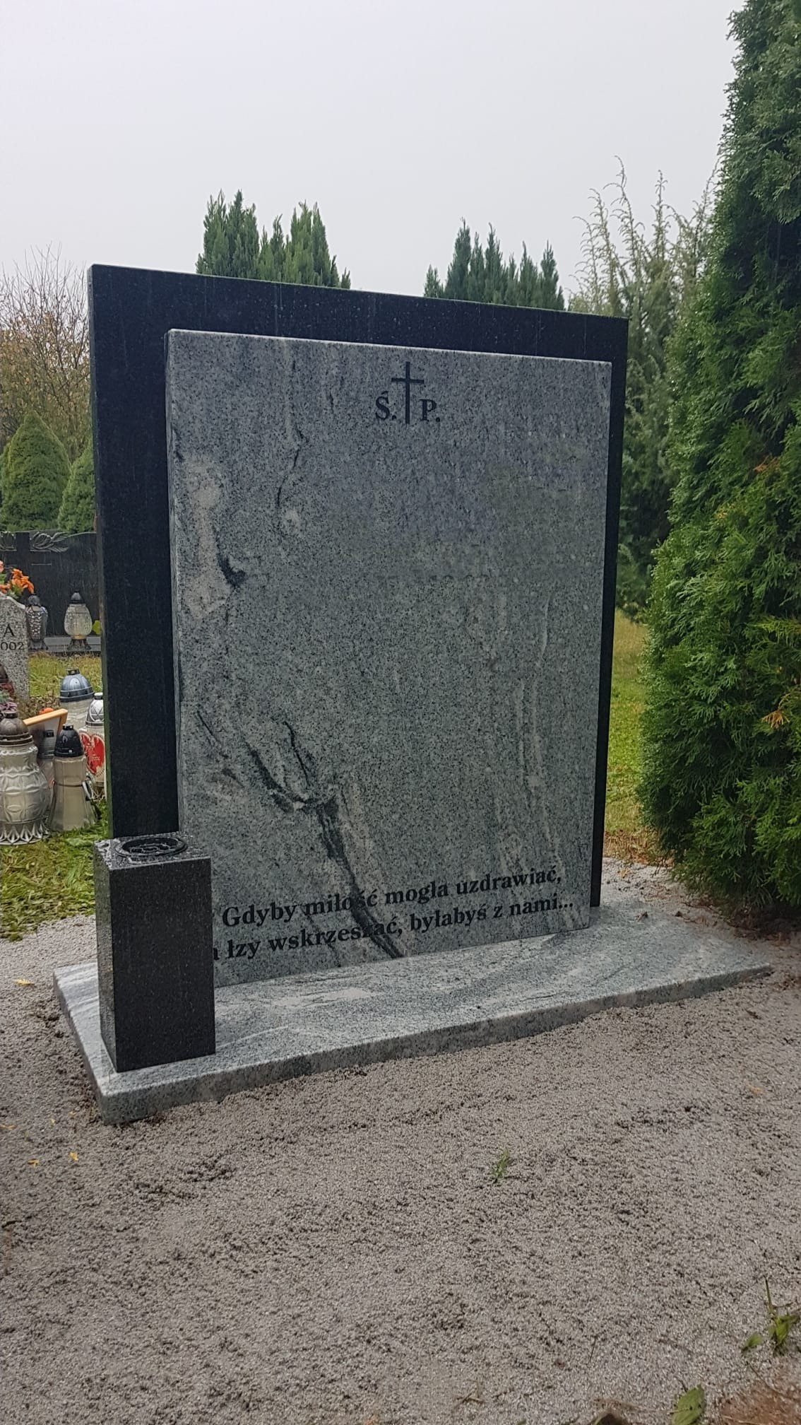 Granitowy pomnik z wygrawerowanym napisem i symbolem krzyża, otoczony zielenią cmentarza.