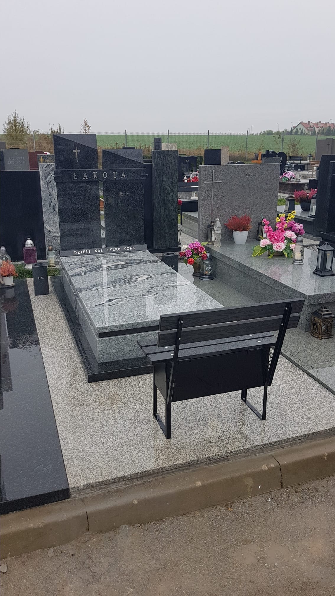 Kompletny nagrobek z jasnoszarego i ciemnoszarego kamienia z napisem 'ŁAKOTA' i ławką z tyłu, otoczony innymi nagrobkami na cmentarzu.