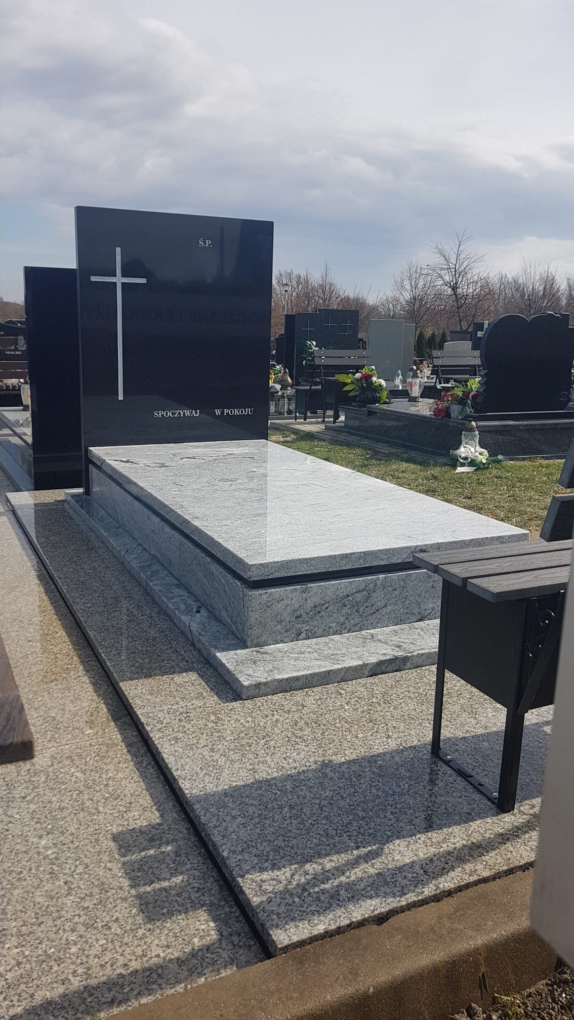 Granitowy pomnik z czarną tablicą, na której widnieje biały krzyż i napis 'S.P. Spoczywaj w pokoju', otoczony innymi nagrobkami na cmentarzu w słoneczny dzień.