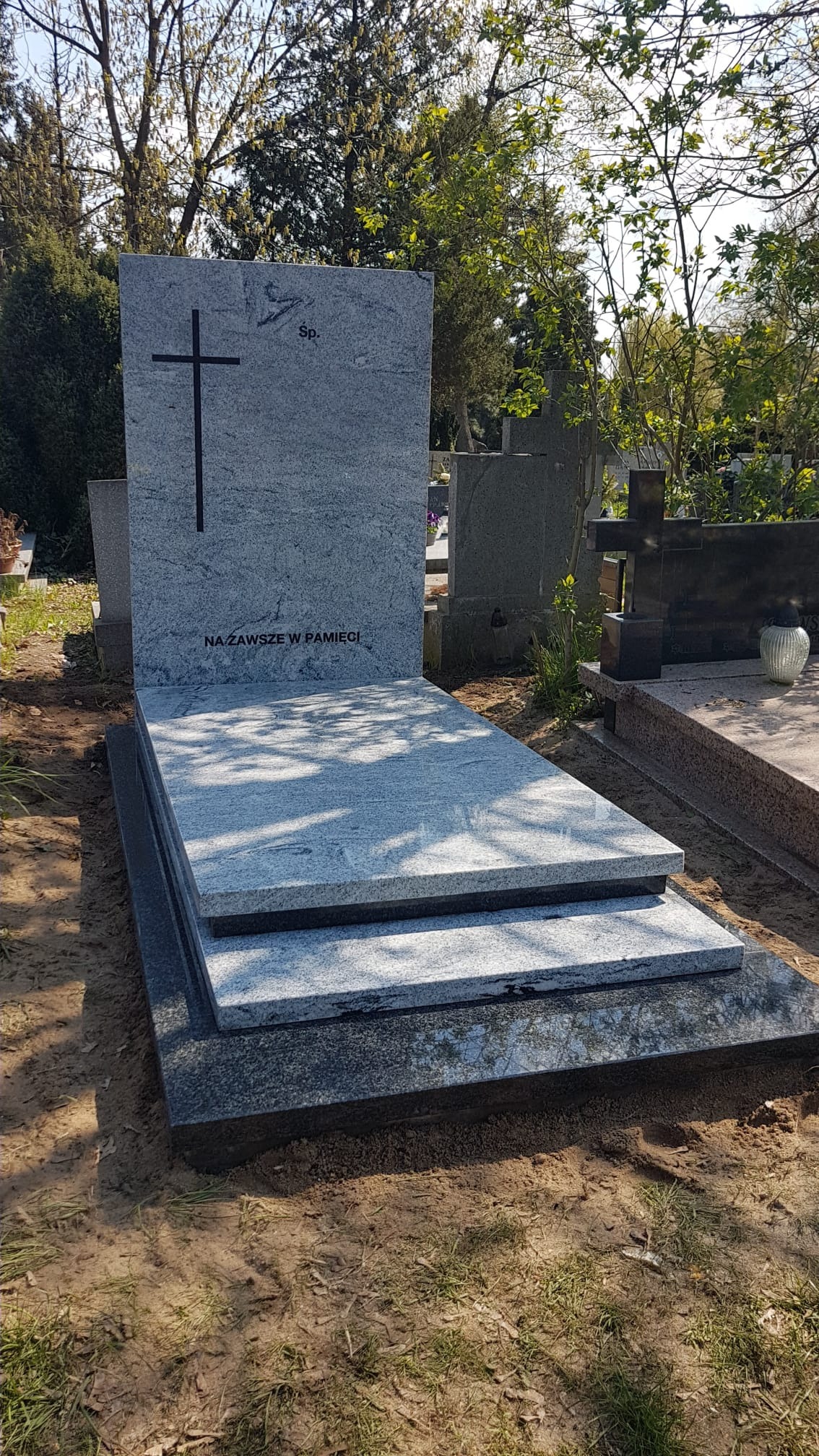 Jasnoszary, nowoczesny pomnik z ciemnoszarymi elementami podstawy, z wygrawerowanym krzyżem i napisem 'Na zawsze w pamięci', na tle cmentarza z drzewami.