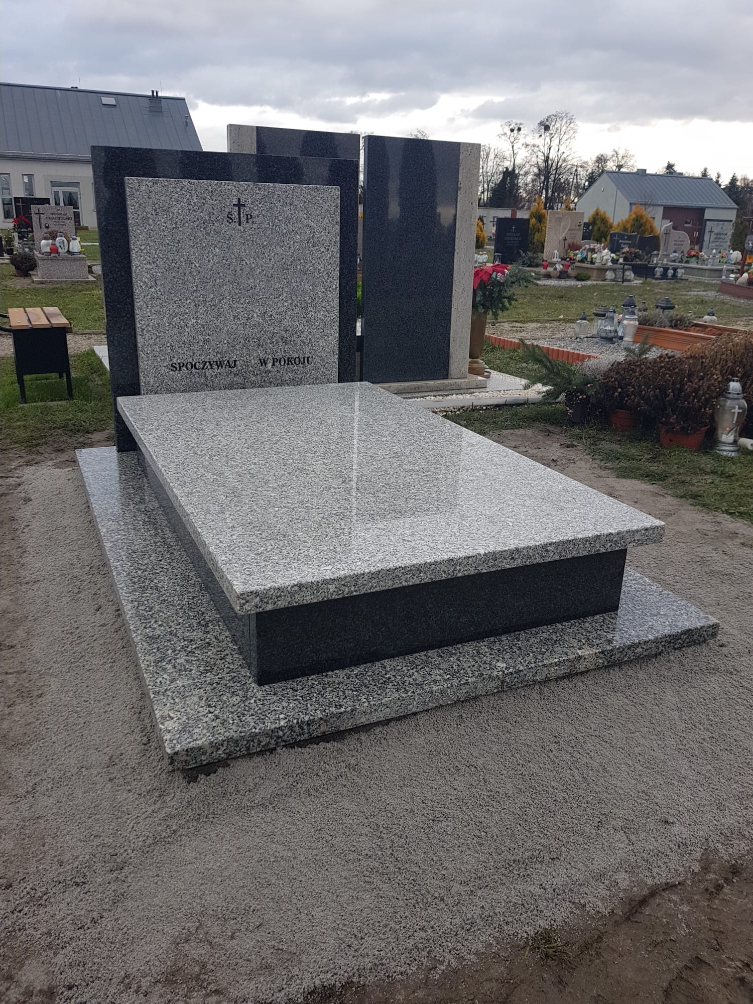 Jasnoszary, dwupoziomowy nagrobek z ciemnoszarym obramowaniem, z napisem 'Spoczywaj w pokoju' i krzyżem, na cmentarzu z widocznymi innymi nagrobkami i budynkami w tle, pochmurne niebo.