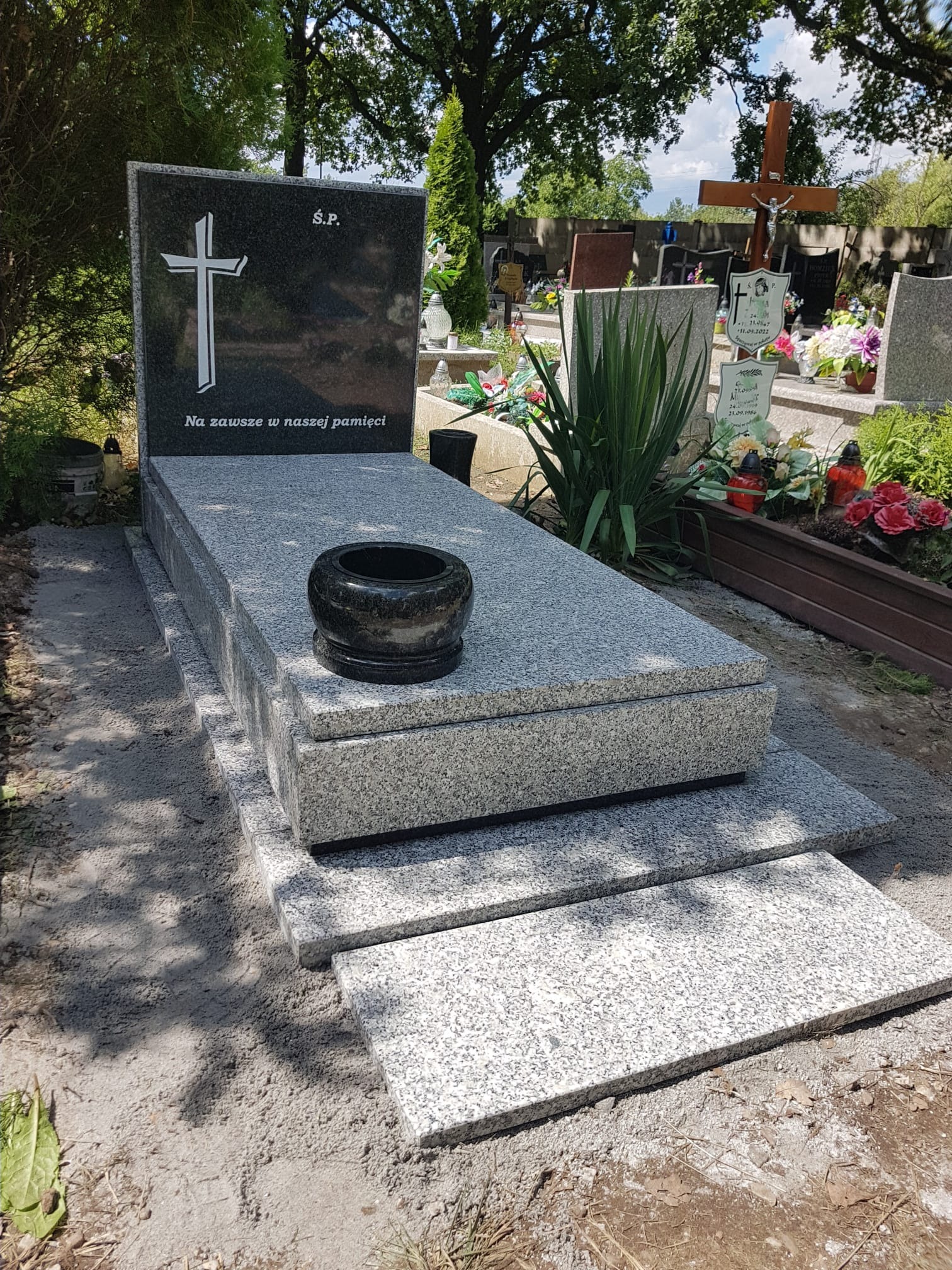 Granitowy pomnik z ciemną tablicą z krzyżem i napisem 'Na zawsze w naszej pamięci', z czarnym wazonem, na tle cmentarza