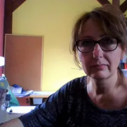 Portret kobiety w okularach, pracującej w Bielawie przy maszynie do szycia, z półkami pełnymi nici w tle.