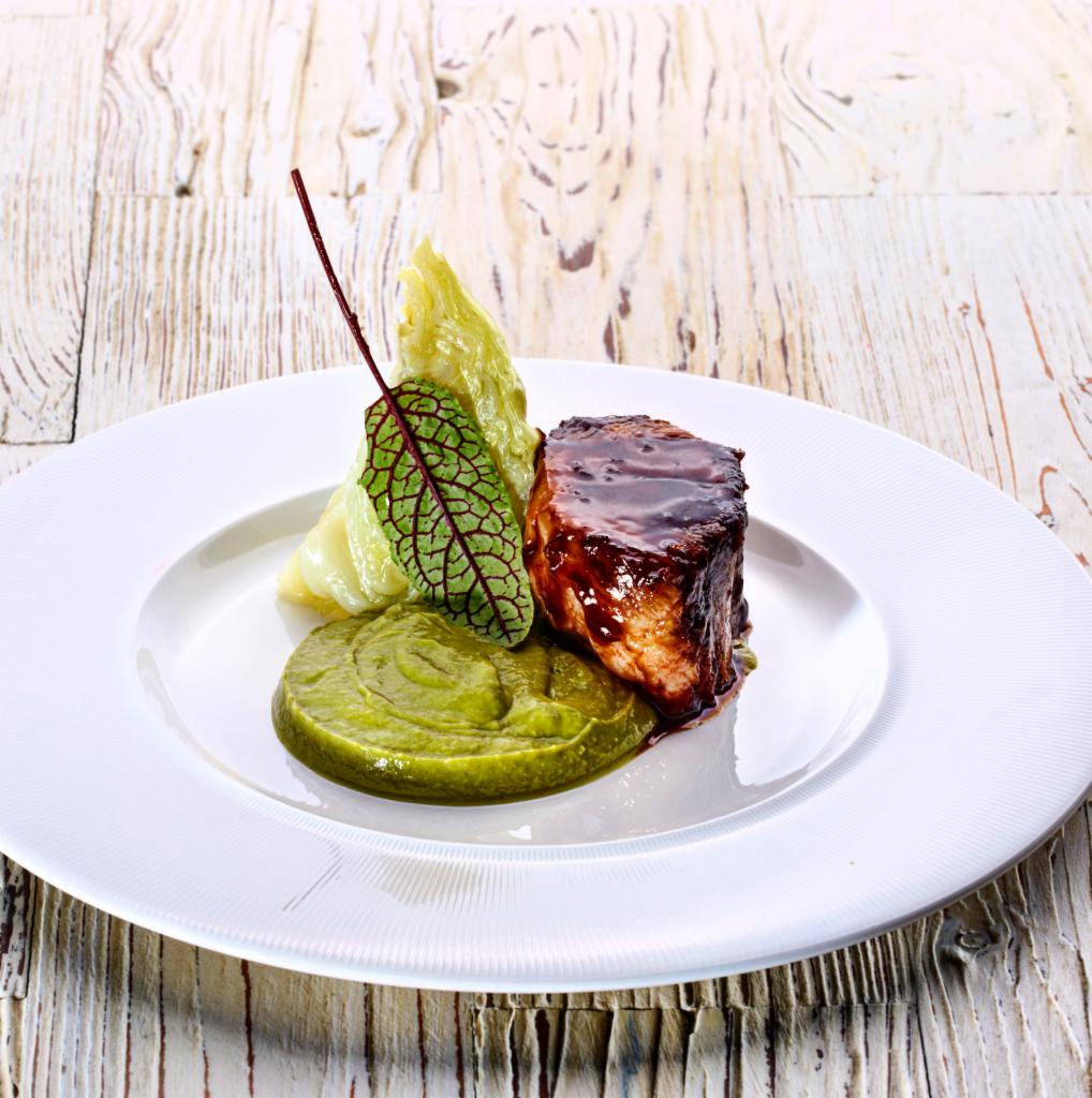 Elegancko podane danie: pieczeń w ciemnym sosie na puree z zielonego groszku, dekorowane liściem i kapustą na białym talerzu na tle drewnianego stołu.