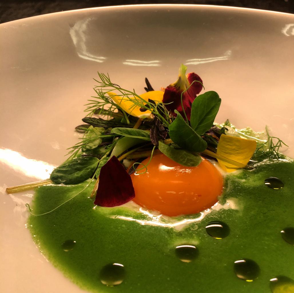 Elegancko podane danie z pomarańczowym żółtkiem, zielonym sosem, świeżymi ziołami i jadalnymi kwiatami na białym talerzu, widoczne krople oliwy.