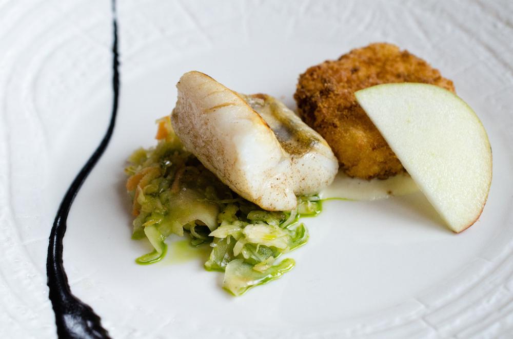 Elegancko podane danie: smażona ryba na zielonej sałatce, obok kotlet i plaster jabłka na białym talerzu z dekoracyjnym sosem.