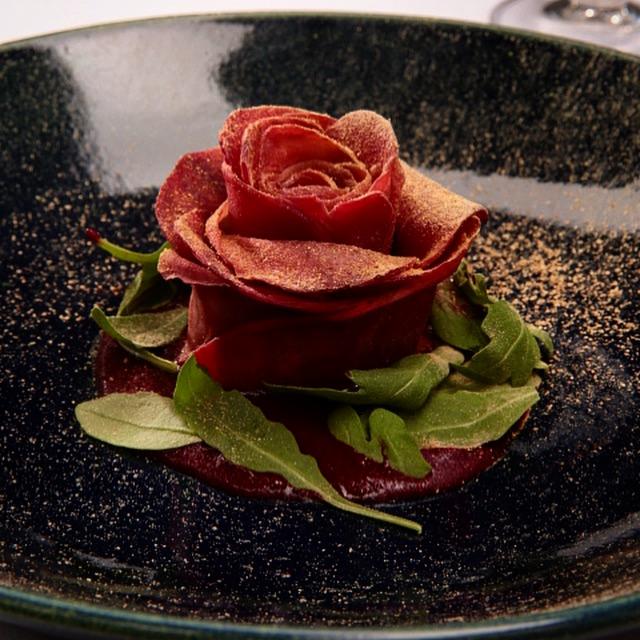 Wykwintna kompozycja z buraka uformowana w kształt róży, otoczona rukolą i posypana złotym pyłem na ciemnym, nakrapianym talerzu.