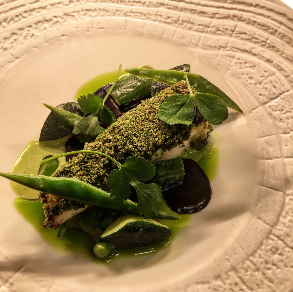 Elegancko podane danie: ryba w zielonej panierce, zielone warzywa, purpurowe plastry, całość polana zielonym sosem na talerzu z tłoczeniami.