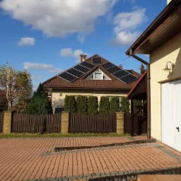 Dom jednorodzinny z brązowym dachem, na którym zamontowane są panele słoneczne, widoczne zza drewnianego płotu i brukowanego podjazdu w słoneczny dzień.