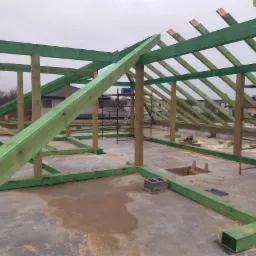 Konstrukcja drewniana dachu w trakcie budowy, impregnowana na zielono, widoczne elementy więźby dachowej, szalunek, bloczki betonowe, w tle domy jednorodzinne.