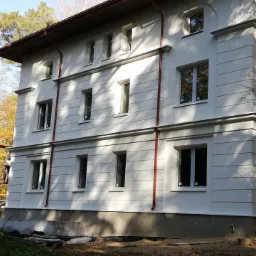 Dwukondygnacyjny dom z jasną elewacją, widoczne poziome pasy dekoracyjne, czerwone rynny, częściowo zasłonięty rusztowaniem, otoczony drzewami w jesiennych kolorach.