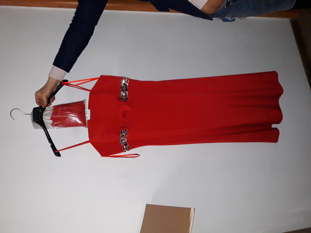 Czerwona, długa suknia wieczorowa z ozdobnymi aplikacjami na wieszaku, częściowo w plastikowym opakowaniu, trzymana przez osobę.