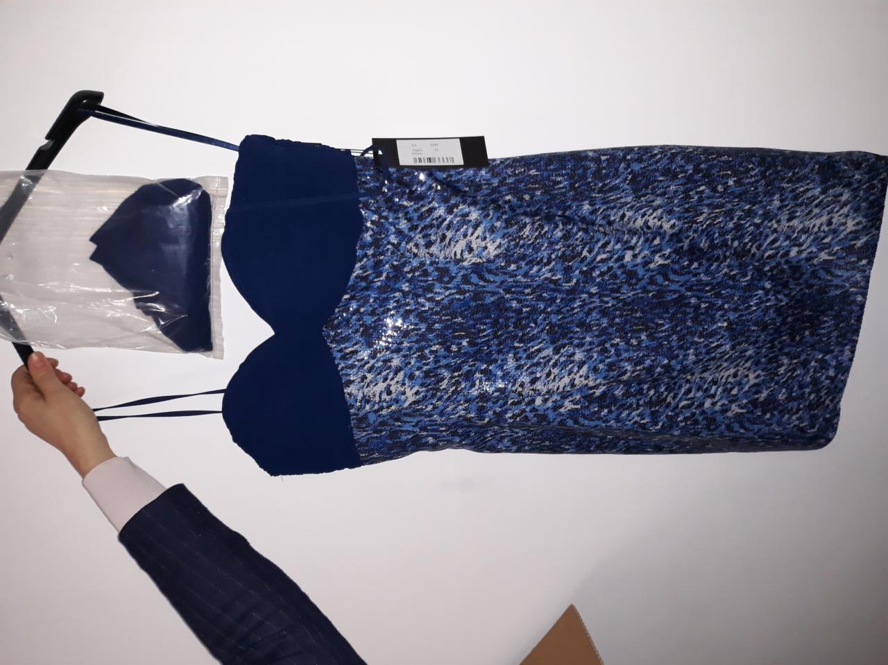 Sukienka damska z niebieskim wzorem cętek i ciemnoniebieską górą, zawieszona na wieszaku obok zapakowanej w folię części garderoby, prawdopodobnie dodatku do sukienki.