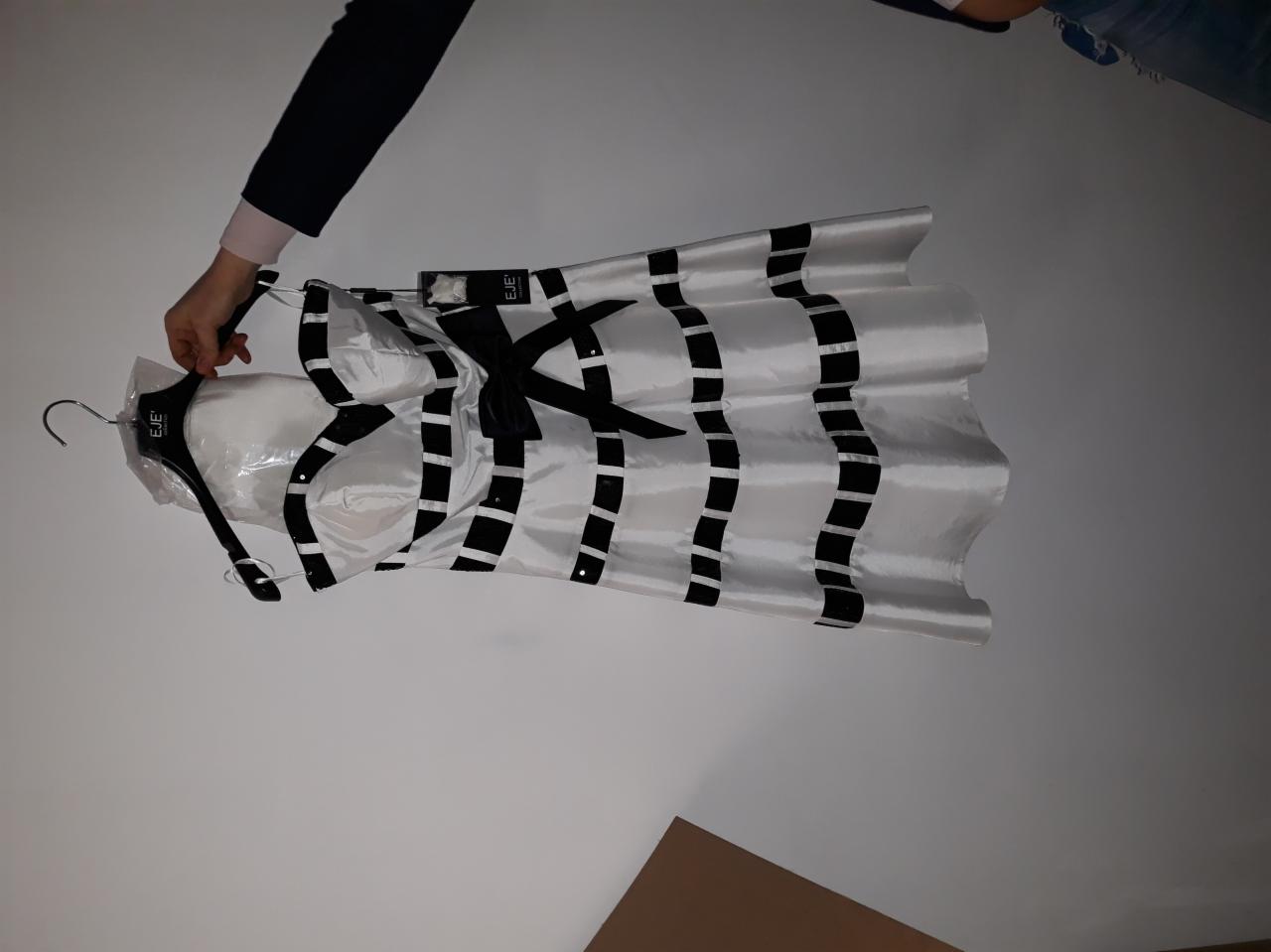 Biała sukienka z czarnymi pasami i kokardą, wisząca na wieszaku, częściowo w folii ochronnej, trzymana przez rękę.