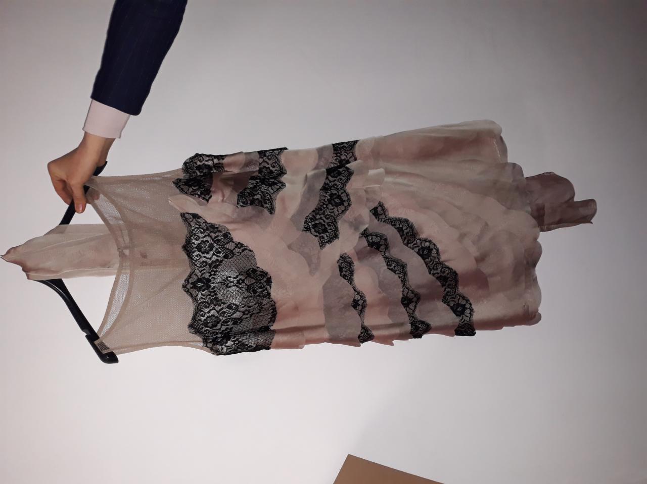 Delikatna, beżowo-różowa sukienka z czarną koronką, zawieszona na wieszaku, trzymana przez osobę w granatowej marynarce.