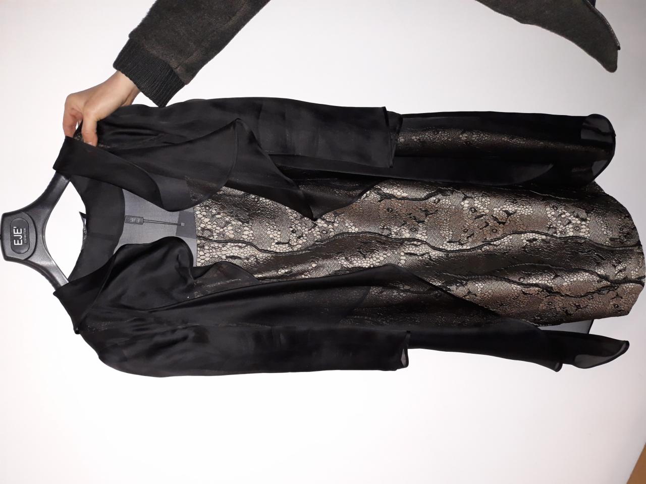 Czarna, satynowa bluzka z koronkową wstawką leży na wieszaku, trzymana przez dłoń w swetrze.