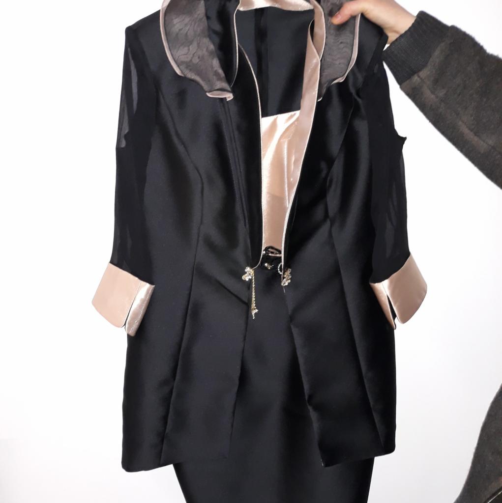 Czarny damski kostium z satynowymi wstawkami w kolorze pudrowego różu, prezentowany na wieszaku, z widoczną ręką w szarym swetrze.