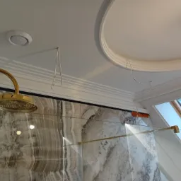 Wykończona łazienka z kabiną prysznicową z marmurowymi płytami, złotą armaturą i oknem dachowym, widoczne niedokończone prace na suficie.